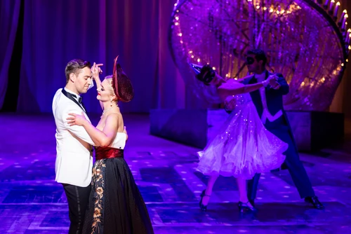 Links im Bild ein junger Mann in weißem Jackett, schwarzer Hose und schwarzer Fliege und eine blonde Frau in schwarz-rotem Kleid und rotem Hut in inniger Tanzpose, rechts im Hintergrund ein auf dem Boden liegender Kronleuchter und ein maskiertes tanzendes Paar in violettem Licht
