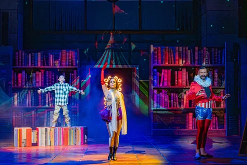 Actors on stage in front of tall bookshelves onto which a circus tent is projected. One person stands on a platform with arms outstretched, two others stand next to them in circus costumes. Scene in bold colours, blue background.