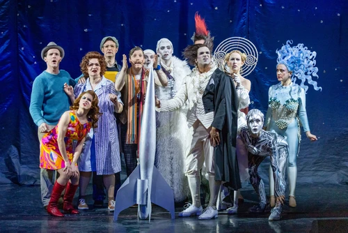 A large group of stage characters stands in front of a dark blue background with a hint of a star pattern. In the centre is a shiny silver rocket prop. The characters wear very different, colourful and decorative costumes, including a colourful, retro-inspired dress, a light fur-like costume, a metallic full-body costume with ornamental headdress, a spiral-shaped head accessory and other outfits with glittering or textured fabrics. They look surprised. Cool, blue-white light.