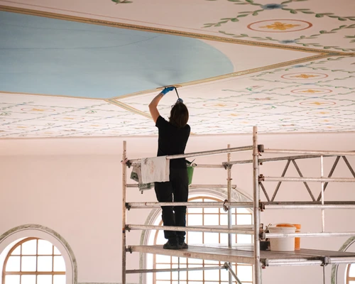 Reconstruction work on the ceiling of the sala thermarum; the decorated ceiling is visible, with a female painter standing on scaffolding 