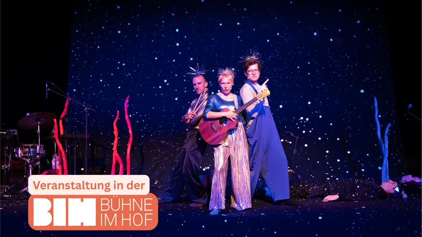 Szenenbild aus "Annas Aquarium und die fetzigen Flundern" von und mit Anna Mabo, mit drei Performer:innen auf der Bühne, teils in Kostümen aus reflektierenden Stoffen.