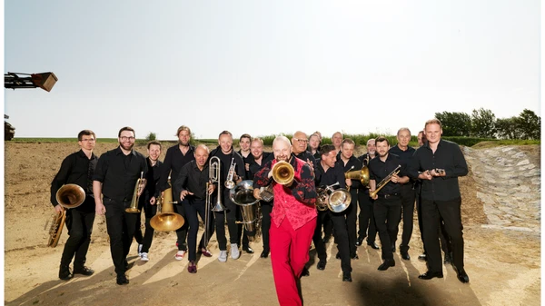 Eine große Gruppe Musiker mit Blasinstrumenten, vorne ein Mann in roter Hose und rotem  Gilet mit Flügelhorn