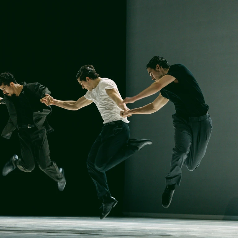 A still from ‘Obsidian’, a choreography by Bobbi Jene Smith and Or Schraiber. The photograph shows dancers from Ballet BC, three of whom are in the foreground, jumping simultaneously, whilst in the background a dancer in a black dress stands against the grey stage backdrop.