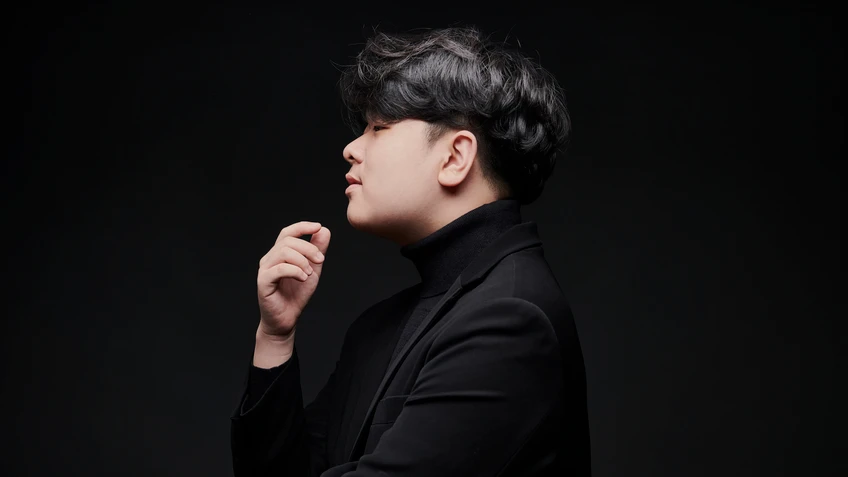 Mann mit dunklem Haar in dunklem Anzug vor schwarzem Hintergrund, die Hand vor seinem Kinn