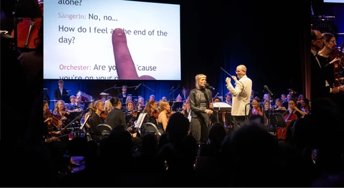 Foto von "Orchesterkaraoke", auf der Bühne ein Orchester, dahinter wird offenbar ein Songtext projiziert, eine Person steht an einem Mikrofon und singt.