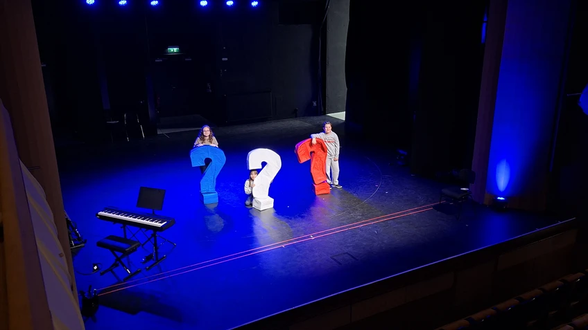 Drei Kinder mit drei großen Fragezeichen auf einer dunkelblau beleuchteten Bühne