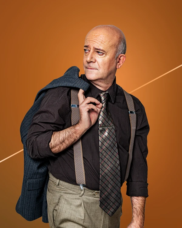 A middle-aged man stands against an orange background, wearing a shirt, tie, suspenders, and a blue blazer over his shoulder.