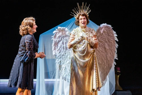 Two people are standing side by side on a theatre stage. One is wearing dark clothes, the other a gold-coloured costume with wings and a radiant headdress. In the background is a piece of furniture covered with a white, transparent cloth.