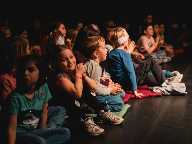The young audience at the BIG BANG Festival sits eagerly on the floor awaiting the next performance. A girl in the front row claps her hands ecstatically, while a boy two seats away from her wears a BIG BANG cardboard crown. 