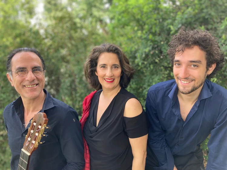 Gruppenfoto Karmon Vounelakos Trio. Drei Personen stehen im Freien mit Bäumen im Hintergrund und lächeln in die Kamera. In der Mitte steht Shira Karmon.