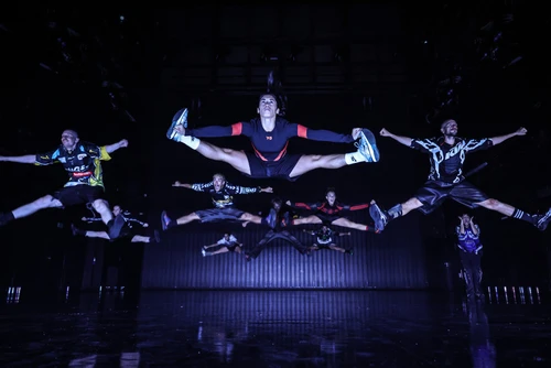 Performancefoto von "Trailer Park", choreografiert von Moritz Ostruschnjak, getanzt von tanz mainz. Die Tänzer:innen in oversized Sporttrikots springen synchron einen Split.