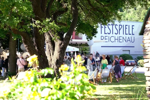 Besucherinnen und Besucher bei sonnigem Wetter im Gartenbereich der Festspiele Reichenau vor dem Festspielgebäude.