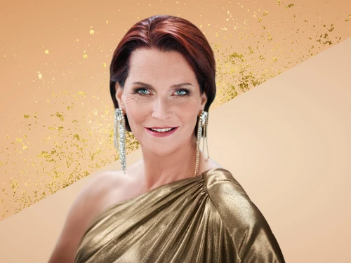 Porträtaufnahmen von Maya Hakvoort in einem eleganten, einseitig drapierten, goldfarbenen Kleid mit auffälligen, langen Ohrringen. Der Hintergrund ist in warmen Beige- und Goldtönen gestaltet, mit dekorativen Glitzerelementen.
