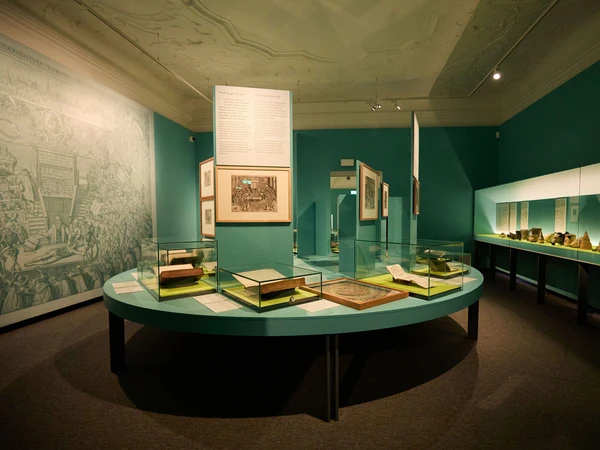 Blauer Raum mit blauem Tisch, darauf sind mehrere Glasvitrinen mit verschiedenen Büchern und Dokumenten. Auf der rechten Wand eine lange Wandvitrine mit verschiedenen Gefäßen.