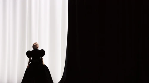 Junge blonde Dame in opulentem schwarzem Kleid vor hellem Vorhang neben schwarzer Wand