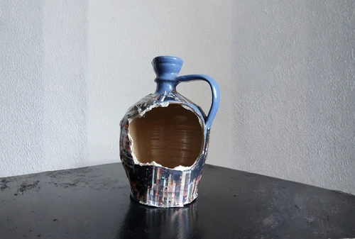 zerbrochener, blauer Krug auf dunklem Tisch vor weißer Wand