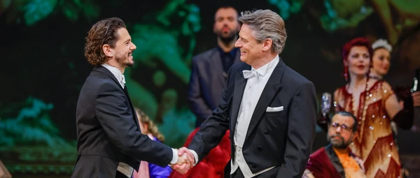 Two men in suits shake hands, in the background there are some people and a red couch
