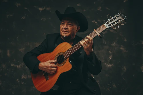 Porträtfoto von Musiker Elias Ochoa mit seinem Instrument, einer Akustikgitarre, gekleidet in einen schwarzen Anzug mit schwarzem Hut auf.