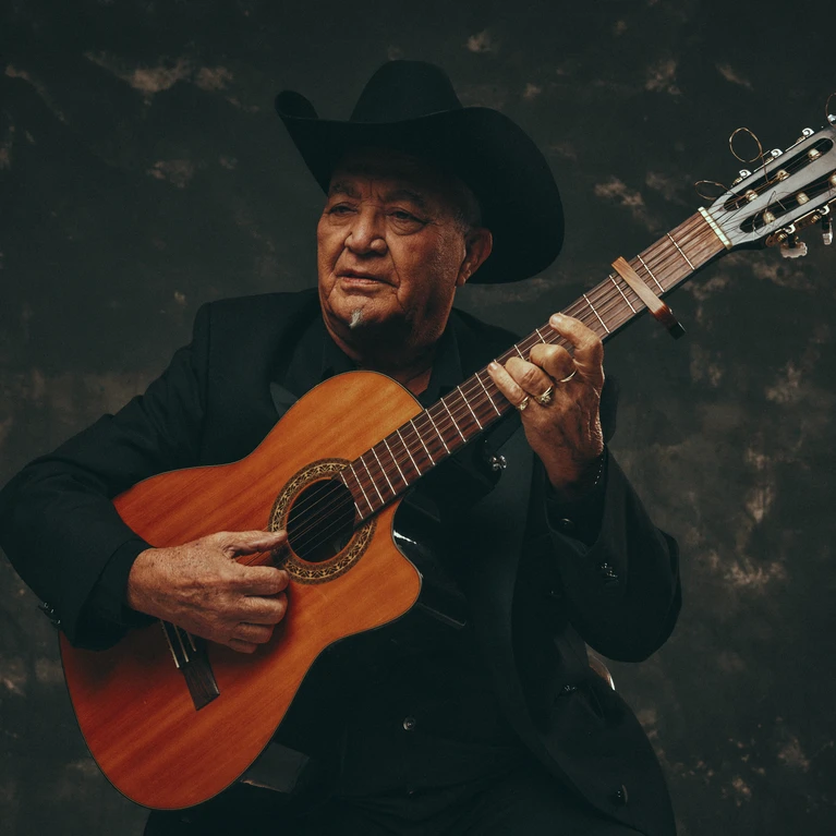 A portrait photograph of musician Elias Ochoa with his instrument, an acoustic guitar, wearing a black suit and a black hat.