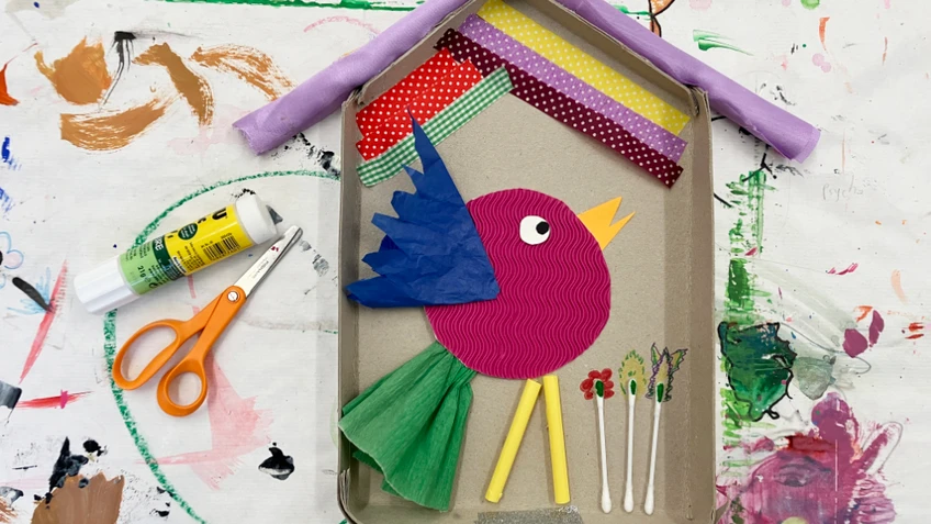 Bunte Bastelarbeit in Form eines vogelförmigen Motivs, gestaltet aus Papier, Stoffresten und kleinen Materialien in einem kartonähnlichen Rahmen. Daneben liegen eine Schere und ein Klebestift auf einer farbverschmierten Unterlage.