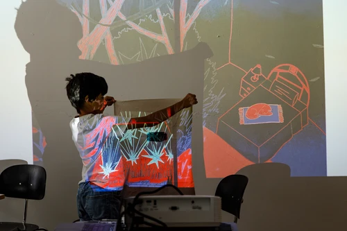 A child holds a cushion cover in front of a wall. The projection of a colourful illustration of a red dog lying on a bed lies on him, the cushion cover and the wall.
