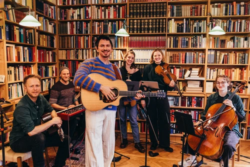 Ein Gitarrist in blau-orange-gestreiftem Pullover in einer kleinen Bibliothek mit 5 MusikerInnen und ihren Instrumenten