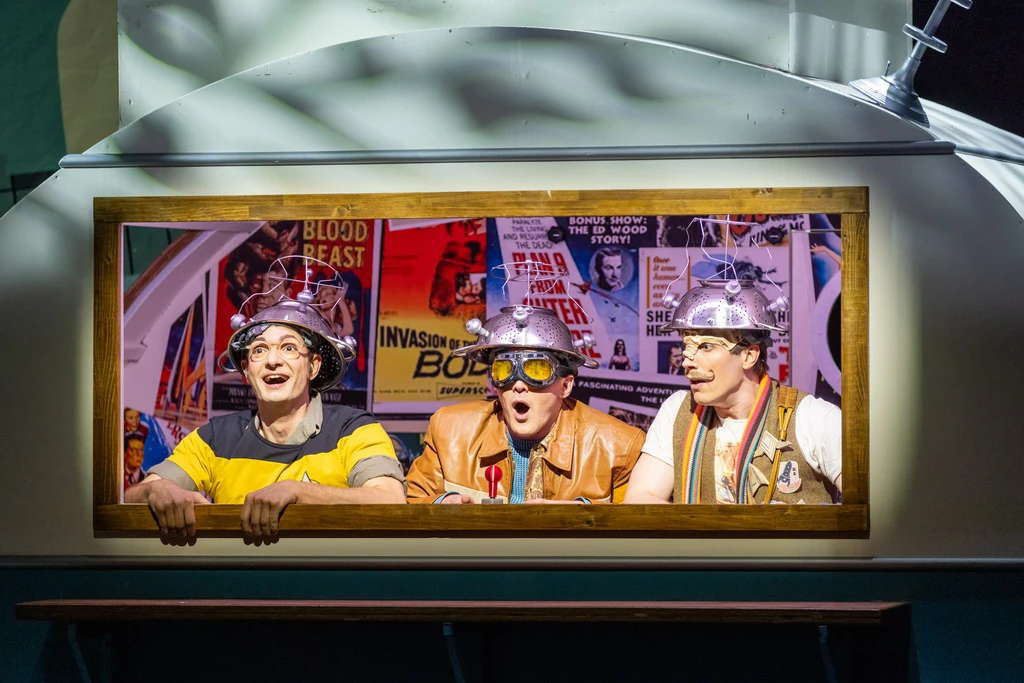 Three people in a well-lit window, wearing improvised helmets on their heads, with old film posters in the background. The expressions on the three men's faces suggest excitement or anticipation. The illuminated frame clearly sets the scene apart from the darker stage background. 