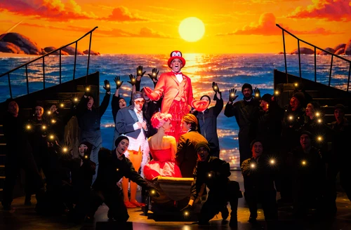 An elaborately staged scene features several performers against a projection of a golden sunset over the sea. At the centre is a person in a bright red costume with a striking hat, standing on a raised platform. In front of them, two people are sitting in a small boat or tub, whilst other performers dressed in black frame the scene and hold up glowing hands. The warm orange and gold tones of the backdrop create a dramatic, fairy-tale atmosphere.
