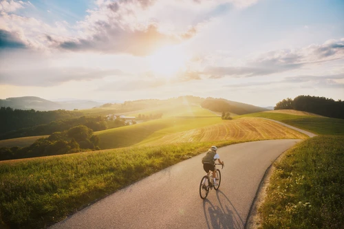 Radfahrer fährt über eine Straße durch idyllische Hügellandschaft, die Sonne scheint