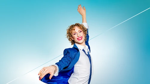 Person in blue suit striking an expressive pose against a blue background; face blurred, red fingernails visible.