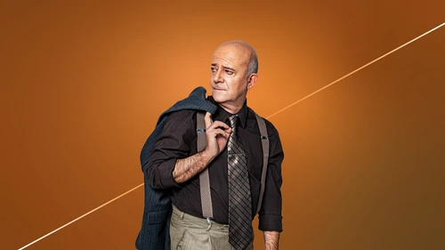 A middle-aged man stands against an orange background, wearing a shirt, tie, suspenders, and a blue blazer over his shoulder.