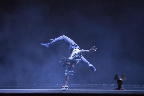 Performancefoto aus dem Tanzstück "Kaléidoscope", choreografiert von Mourad Merzouki, getanzt von der Compagnie Käfig. Ein Tänzer tanzt eine akrobatische Figur, bei der er lediglich mit einer Hand den Boden berührt, der Rest seines Körpers ist in der Luft, ein Bei nach hinten im Sprung abgewinkelt. Die Bühne ist in blaues Licht und durchsichtigen Bühnennebel gehüllt.