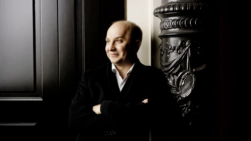 Mann in Anzug lehnt an einer prunkvollen dunklen Säule vor einem großen Tor 