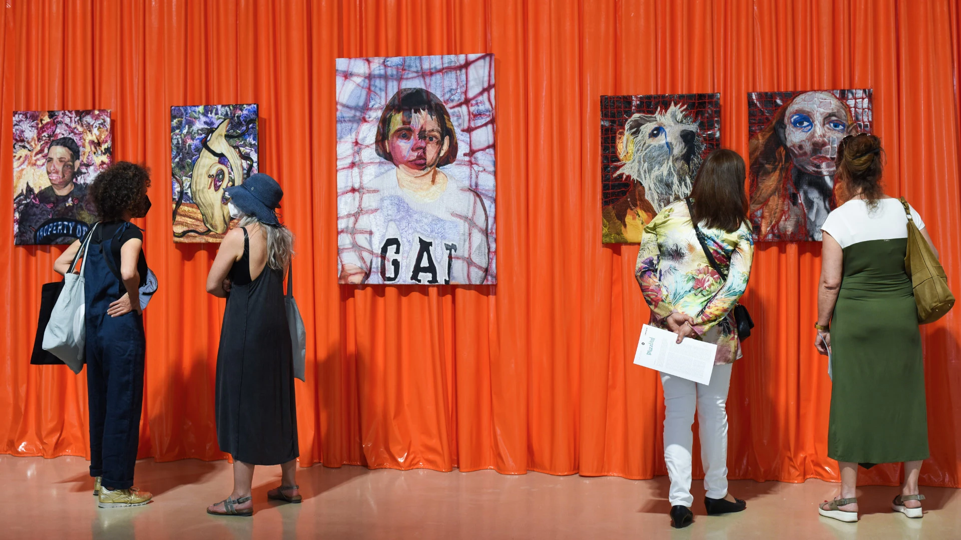Besucherinnen betrachten farbintensive Porträtgemälde vor leuchtend orangefarbenem Hintergrund in einer Galerie.