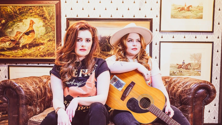 Cassie und Maggie sitzen lässig auf einem braunen Ledersofa in einem rustikal dekorierten Raum mit Jagdtrophäen und Gemälden, wobei eine eine Gitarre hält und beide direkt und selbstbewusst in die Kamera blicken.