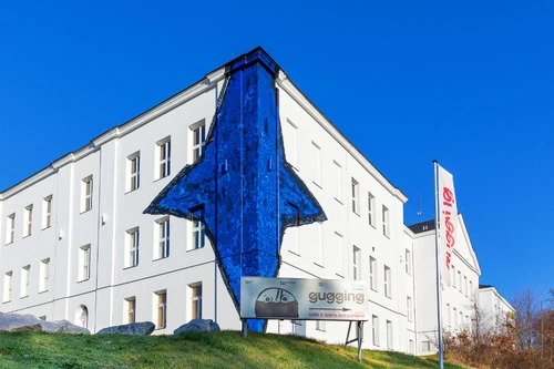 Weißes Museumsgebäude mit auffälligem blauen Wandgemälde in Pfeilform und Beschilderung „Gugging“.