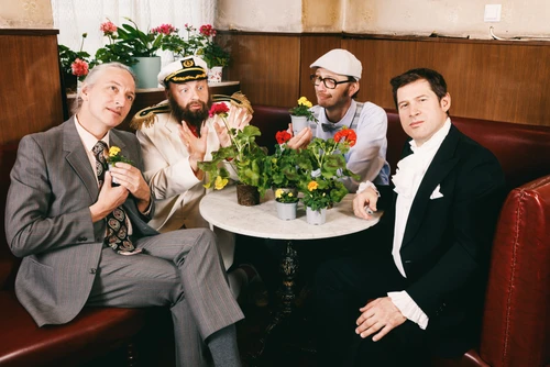 Vier Männer sitzen um einen Kaffeehaustisch auf einer roten Lederbank, auf dem Tisch stehen Blumen in Töpfen. Ein Mann trägt ein Kapitänskostüm