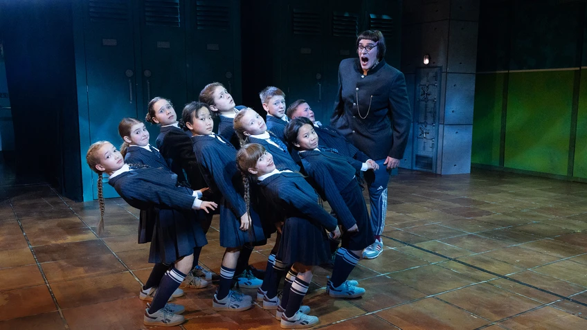 Gruppe von Kindern in dunklen Schuluniformen, weichen erschrocken vor einer großen Person zurück, von der sie angebrüllt werden. Fliesenboden, Spinde im Hintergrund, Lichtstimmung kühl und dramatisch.