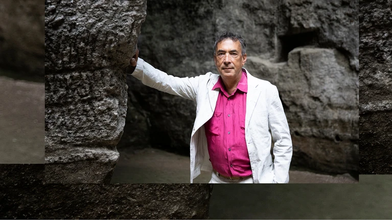 Photo of Hubert von Goisern, wearing a pink shirt, a white suit jacket, and suit pants. He is leaning against a stone wall with one hand; a stone wall can also be seen in the background.