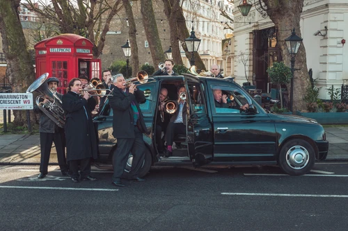 Foto vom London Brass Ensemble mit ihren Instrumenten in und um ein schwarzes Londoner-Taxi.