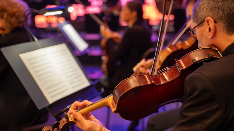 A photo of the Tonkünstler Orchestra performing on a large stage, bathed in pink light. The camera looks over the shoulder of a violinist as he plays; his music stand, with the score open, is also visible in the background.