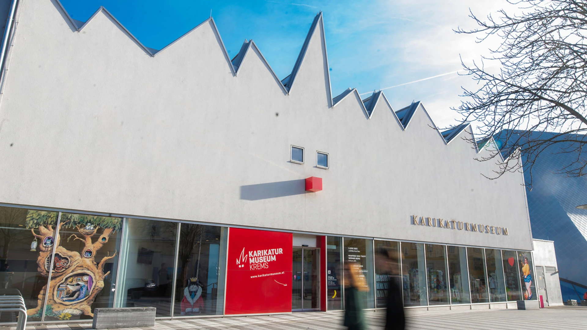 Moderne Museumsfassade mit gezacktem Dach und großem roten Eingangselement, davor Passanten.