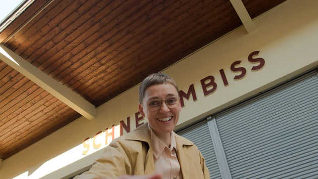 A portrait photograph of artist and chef Steffi Parlow, taken from a low angle; behind her, the word ‘Imbiss’ is visible on a shop front; she is in the foreground, smiling at the camera and pointing with one hand.