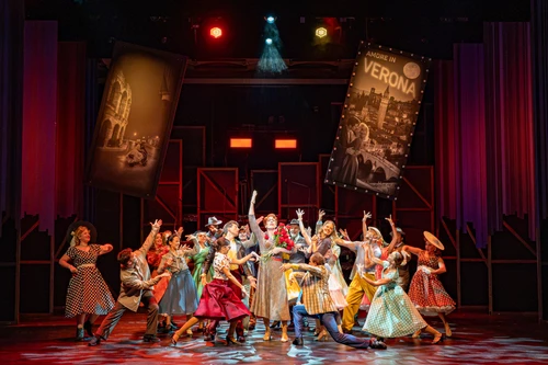 A large ensemble scene on a theatre stage featuring numerous performers gathered in a tight group at the centre of the stage. Many of the performers have their arms stretched upwards or are striking dynamic poses. The stage is illuminated by warm red and orange lighting, as well as occasional blue highlights. In the background, two large banners or posters hang diagonally above the scene, whilst tall curtains and other stage elements frame the space. The patterned stage floor enhances the effect of the central choreography. The composition draws the eye to the group’s collective movement and conveys the impression of a festive or concluding ensemble performance.

