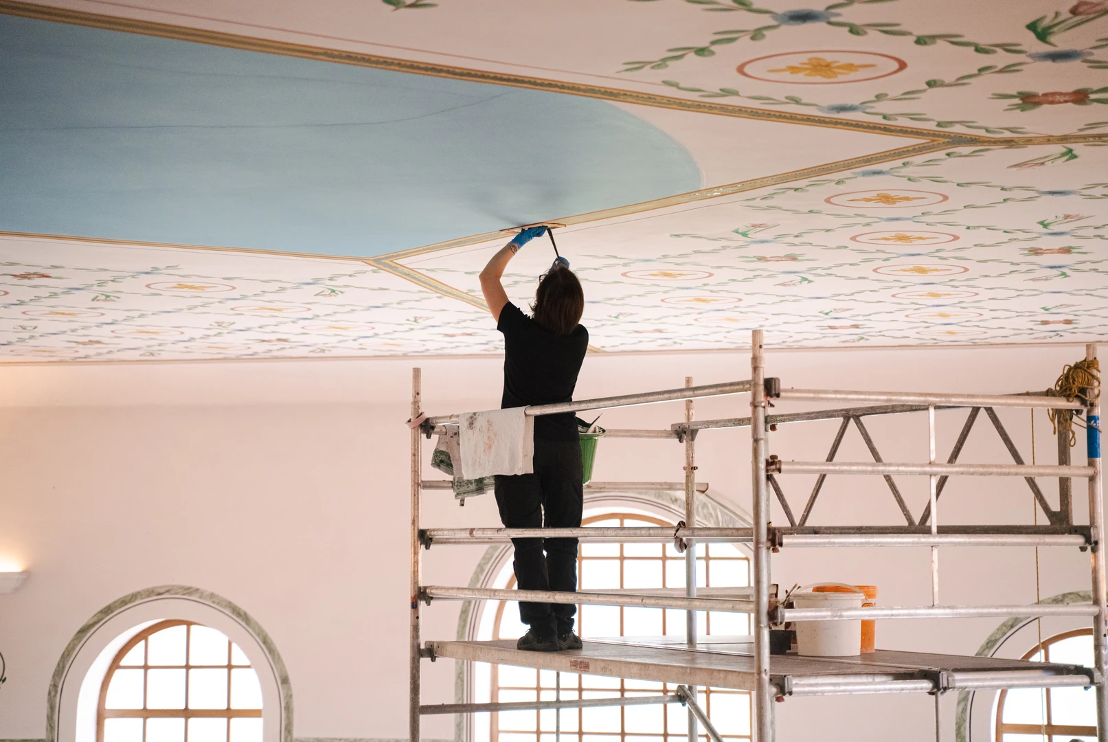 Reconstruction work on the ceiling of the sala thermarum; the decorated ceiling is visible, with a female painter standing on scaffolding 