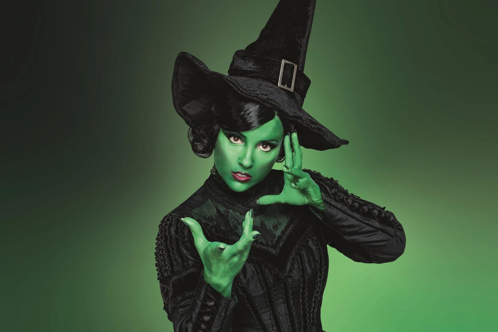 Young woman with green skin and black hair, dressed in black, wearing a pointed black witch's hat, looking at the camera, hands in a dramatic pose, green background