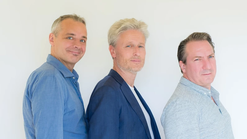 Thomas Maurer, Florian Scheuba und Robert Palfrader stehen seitlich versetzt hintereinander vor hellem Hintergrund, tragen blaue Hemden und Jacken und blicken mit leicht unterschiedlichen, ruhigen Gesichtsausdrücken in die Kamera.