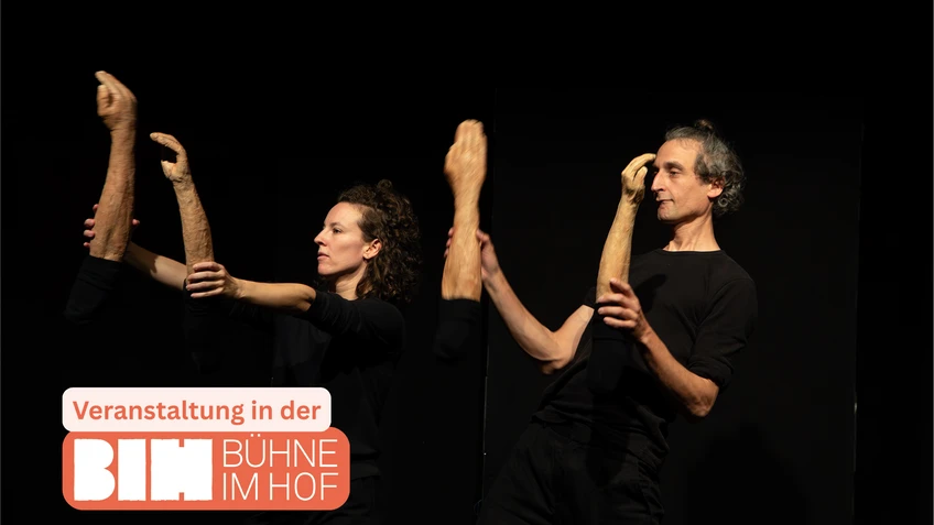 Foto vom "Zirkus Anschauen!", zwei Performer:innen in schwarzen T-Shirts stehen vor einem komplett schwarzen Bühnenhintergrund. Sie halten modellierte, naturfarbene Unterarme inklusive Händen hoch.