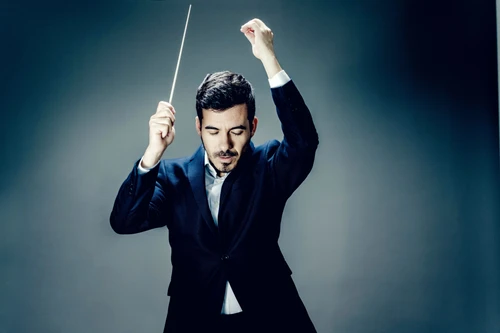 Foto von Dirigent Nuno Coelho, gekleidet in einen blauen Blazer mit am Kragen leicht geöffnetem hellblauem Hemd. Zu sehen ist er vor einem einfarbigen blau-grauen Hintergrund, wie er dirigiert, den Taktstock in einer Hand.