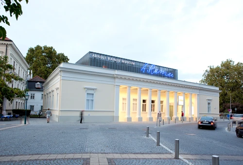 Außenansicht des Museumsgebäudes mit historischer Fassade und Säulengang vor blauem Himmel davor hellgrauer Straßenzug